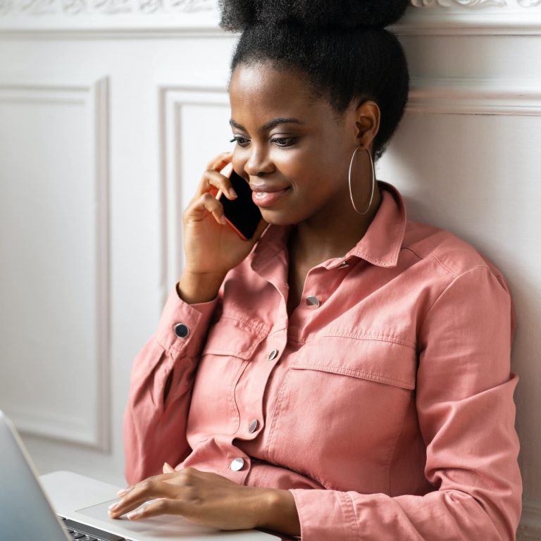 African American millennial woman talking on cellphone, working on laptop at home